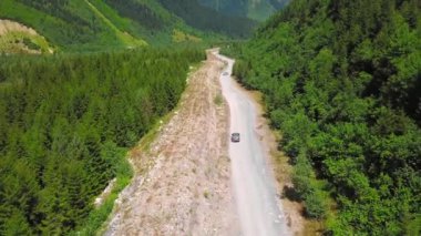 Araba vadilerdeki dağların arasında tozlu yol boyunca ilerliyor. Dağ nehri yakınlarda akar. Dağlarda arabayla ve kırsal kesimde off-road ile seyahat etmek. İnsansız hava aracı görüntüsü. Güneşli yaz gününde yolculuk.