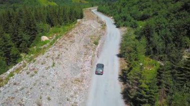 Araba vadilerdeki dağların arasında tozlu yol boyunca ilerliyor. Dağ nehri yakınlarda akar. Dağlarda arabayla ve kırsal kesimde off-road ile seyahat etmek. İnsansız hava aracı görüntüsü. Güneşli yaz gününde yolculuk.