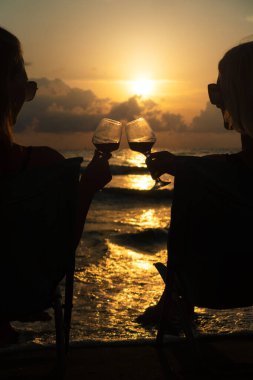 Güneş gözlüklü ve bikinili iki seksi kadının silueti deniz kıyısında katlanan sandalyelere oturmuş ve büyüleyici bir günbatımının arka planına kadeh tokuşturarak şarap içiyorlar.