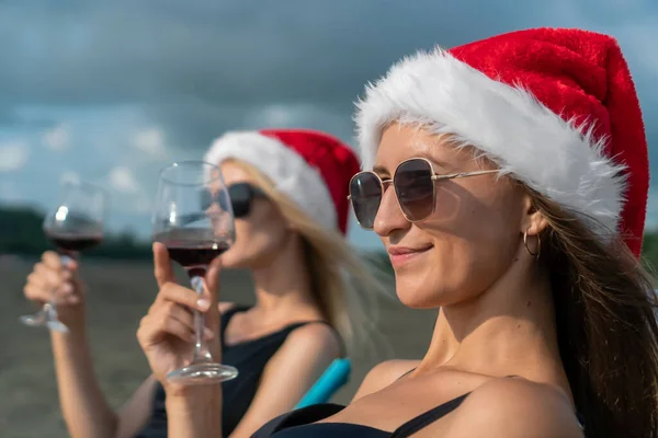 Portrait of two young women in swimsuits, sunglasses and Santa Claus hats with a glass of wine in their hands sitting on folding chairs on the sandy beach of the sea and enjoying the moment.
