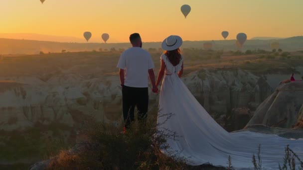 若いカップルが山の上に立って 気球が空を飛ぶのを見る トルコのカパドキア 長いヴィンテージのドレスを地面に伸ばし 白い帽子の女性 手を握ってる 素晴らしい日の出だ — ストック動画