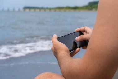 İnternette sörf yaparken siyah ekranlı akıllı telefon kullanan bir adamın yakın plan görüntüsü. Plaj iskelesinde duran 4G internet bağlantısını kullanarak cep telefonu cihazı tutan bir adamın elleri.