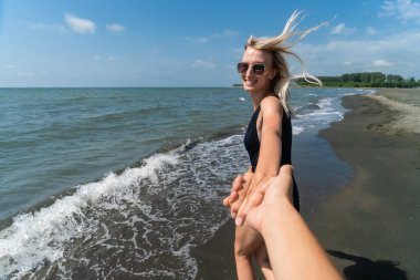 Genç, güler yüzlü, narin, siyah mayo giymiş, güneş gözlüklü bir kadın güneşli bir günde bir adamın elini tutuyor ve deniz kıyısında koşuyor. Beni izleyin.