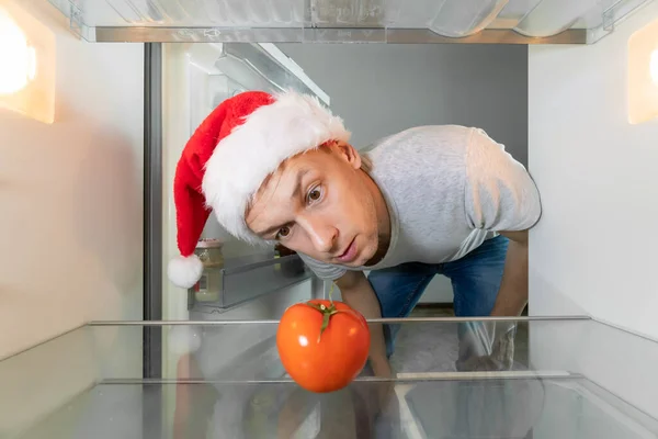 Man in a Santa Claus hat looks suspiciously at a tomato in an empty refrigerator. Concept of an empty refrigerator, delivery service, hunger. Camera inside the refrigerator