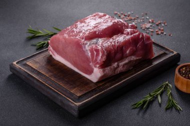Kesme tahtasındaki çiğ domuz bifteği, otlar ve baharatlar koyu taş masada.