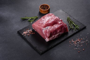 Kesme tahtasındaki çiğ domuz bifteği, otlar ve baharatlar koyu taş masada.