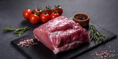 Kesme tahtasındaki çiğ domuz bifteği, otlar ve baharatlar koyu taş masada.