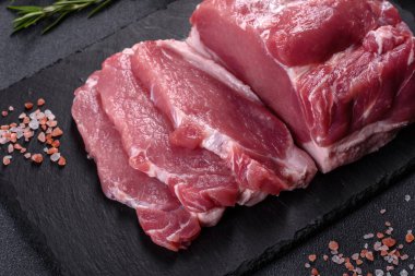 Kesme tahtasındaki çiğ domuz bifteği, otlar ve baharatlar koyu taş masada.