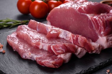 Kesme tahtasındaki çiğ domuz bifteği, otlar ve baharatlar koyu taş masada.