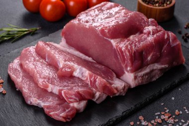 Kesme tahtasındaki çiğ domuz bifteği, otlar ve baharatlar koyu taş masada.