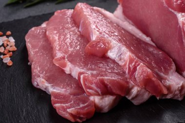 Kesme tahtasındaki çiğ domuz bifteği, otlar ve baharatlar koyu taş masada.