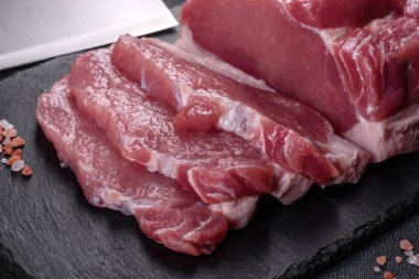 Kesme tahtasındaki çiğ domuz bifteği, otlar ve baharatlar koyu taş masada.