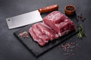 Kesme tahtasındaki çiğ domuz bifteği, otlar ve baharatlar koyu taş masada.