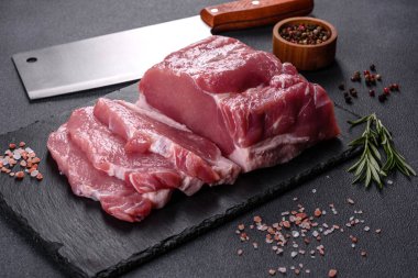 Kesme tahtasındaki çiğ domuz bifteği, otlar ve baharatlar koyu taş masada.