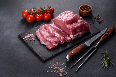 Kesme tahtasındaki çiğ domuz bifteği, otlar ve baharatlar koyu taş masada.