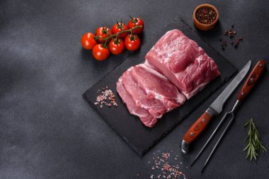 Kesme tahtasındaki çiğ domuz bifteği, otlar ve baharatlar koyu taş masada.