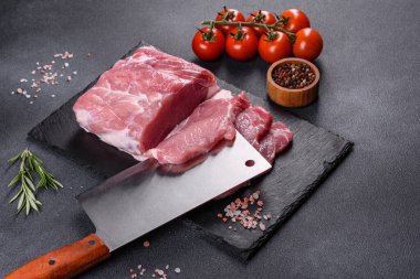 Kesme tahtasındaki çiğ domuz bifteği, otlar ve baharatlar koyu taş masada.