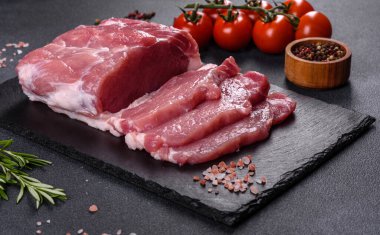 Kesme tahtasındaki çiğ domuz bifteği, otlar ve baharatlar koyu taş masada.