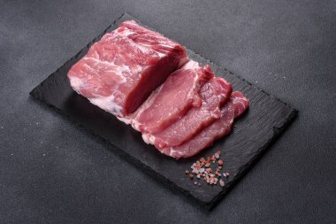 Kesme tahtasındaki çiğ domuz bifteği, otlar ve baharatlar koyu taş masada.