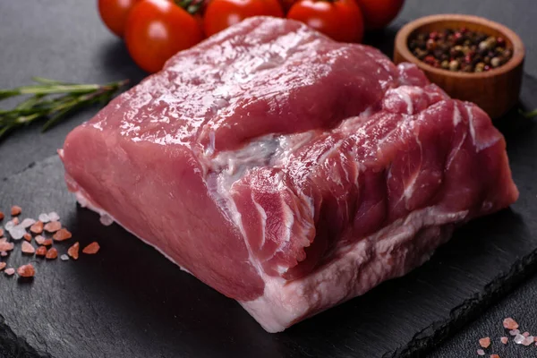 Kesme tahtasındaki çiğ domuz bifteği, otlar ve baharatlar koyu taş masada.