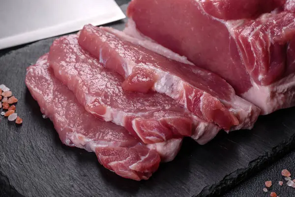 Kesme tahtasındaki çiğ domuz bifteği, otlar ve baharatlar koyu taş masada.