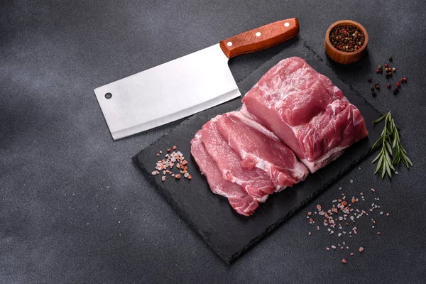 Kesme tahtasındaki çiğ domuz bifteği, otlar ve baharatlar koyu taş masada.