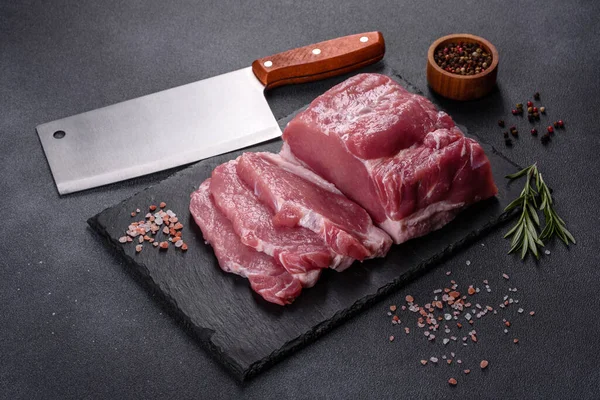 Kesme tahtasındaki çiğ domuz bifteği, otlar ve baharatlar koyu taş masada.