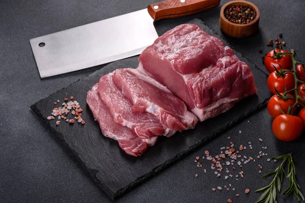 Kesme tahtasındaki çiğ domuz bifteği, otlar ve baharatlar koyu taş masada.
