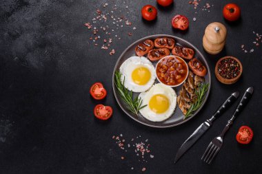 Tam İngiliz kahvaltısı. Fasulye, sahanda yumurta, kızarmış sosis, domates, mantar. Koyu beton bir masada kızarmış ekmek.