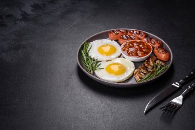 Tam İngiliz kahvaltısı. Fasulye, sahanda yumurta, kızarmış sosis, domates, mantar. Koyu beton bir masada kızarmış ekmek.