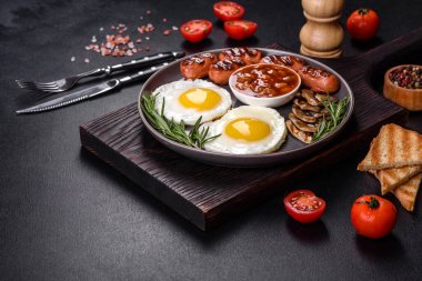 Tam İngiliz kahvaltısı. Fasulye, sahanda yumurta, kızarmış sosis, domates, mantar. Koyu beton bir masada kızarmış ekmek.