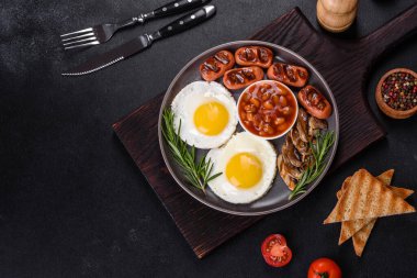 Tam İngiliz kahvaltısı. Fasulye, sahanda yumurta, kızarmış sosis, domates, mantar. Koyu beton bir masada kızarmış ekmek.