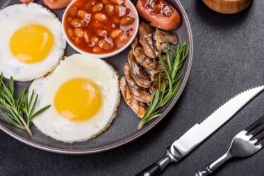 Tam İngiliz kahvaltısı. Fasulye, sahanda yumurta, kızarmış sosis, domates, mantar. Koyu beton bir masada kızarmış ekmek.