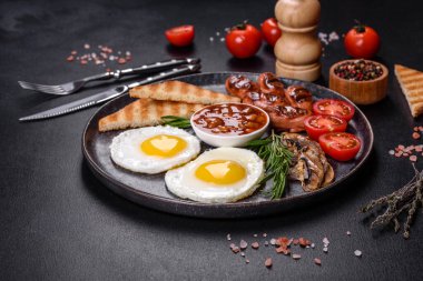 Tam İngiliz kahvaltısı. Fasulye, sahanda yumurta, kızarmış sosis, domates, mantar. Koyu beton bir masada kızarmış ekmek.