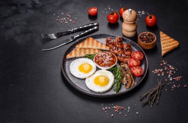 Tam İngiliz kahvaltısı. Fasulye, sahanda yumurta, kızarmış sosis, domates, mantar. Koyu beton bir masada kızarmış ekmek.