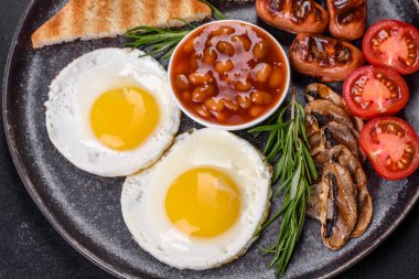 Tam İngiliz kahvaltısı. Fasulye, sahanda yumurta, kızarmış sosis, domates, mantar. Koyu beton bir masada kızarmış ekmek.