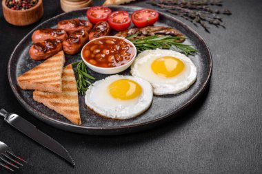 Tam İngiliz kahvaltısı. Fasulye, sahanda yumurta, kızarmış sosis, domates, mantar. Koyu beton bir masada kızarmış ekmek.