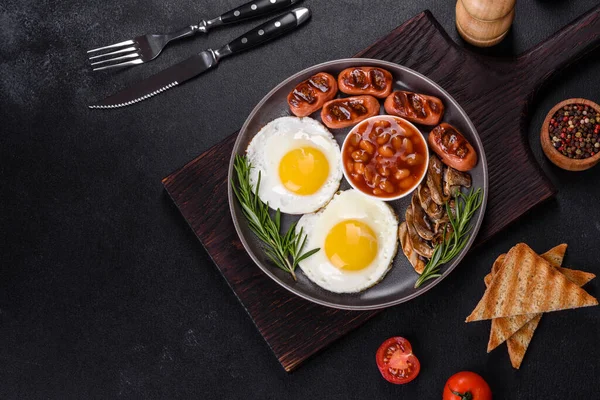 Tam İngiliz kahvaltısı. Fasulye, sahanda yumurta, kızarmış sosis, domates, mantar. Koyu beton bir masada kızarmış ekmek.