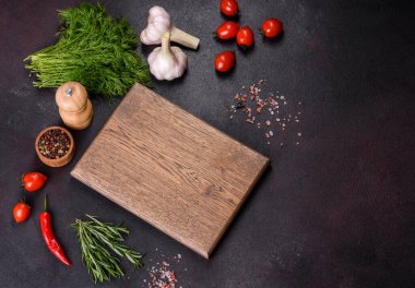 A wooden cutting board with spices, herbs, cherry tomatoes and salt on a black concrete background. Cooking at home