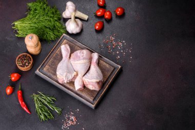 Ahşap bir kesme tahtasının üzerindeki baharatlı ve otlu çiğ tavuğun üç bacağı koyu beton arka planda. Evde yemek pişirmek.