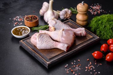 Ahşap bir kesme tahtasının üzerindeki baharatlı ve otlu çiğ tavuğun üç bacağı koyu beton arka planda. Evde yemek pişirmek.