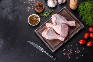 Ahşap bir kesme tahtasının üzerindeki baharatlı ve otlu çiğ tavuğun üç bacağı koyu beton arka planda. Evde yemek pişirmek.