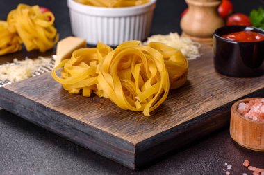 Çiğ tagliatelle macunu, rendelenmiş peynir, kiraz domatesleri, baharatlar ve otlar ahşap kesim tahtasının üzerinde koyu beton bir arka planda.