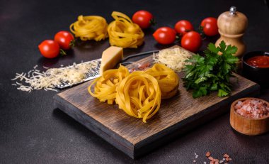 Çiğ tagliatelle macunu, rendelenmiş peynir, kiraz domatesleri, baharatlar ve otlar ahşap kesim tahtasının üzerinde koyu beton bir arka planda.