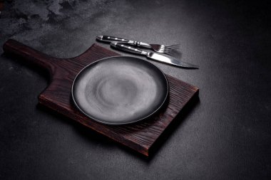 İçinde bıçak, çatal ya da kaşık olan boş bir tabak. Koyu beton arka planda tahta kesim tahtası var. Ev aletleri ve ev yemeği malzemelerinin hazırlanması