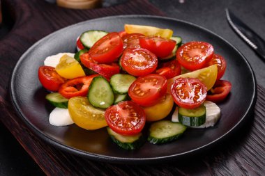 Fresh, delicious salad with cherry tomatoes, cucumbers, sweet peppers, cheese and olive oil on a black plate against a dark concrete background. Vegetarian dish