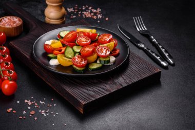 Fresh, delicious salad with cherry tomatoes, cucumbers, sweet peppers, cheese and olive oil on a black plate against a dark concrete background. Vegetarian dish