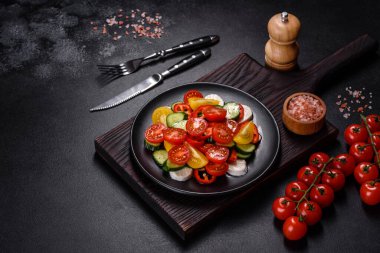 Fresh, delicious salad with cherry tomatoes, cucumbers, sweet peppers, cheese and olive oil on a black plate against a dark concrete background. Vegetarian dish