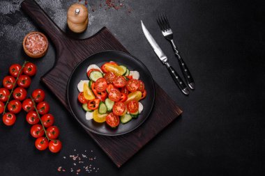 Fresh, delicious salad with cherry tomatoes, cucumbers, sweet peppers, cheese and olive oil on a black plate against a dark concrete background. Vegetarian dish