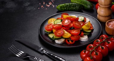 Fresh, delicious salad with cherry tomatoes, cucumbers, sweet peppers, cheese and olive oil on a black plate against a dark concrete background. Vegetarian dish